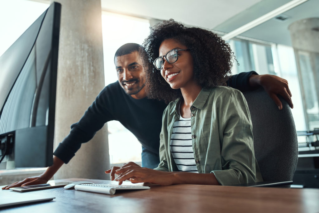 An african young businesswoman sitting at her desk working on computer with male colleague standing by